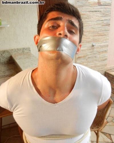 A muscular young man is gagged with silver duct tape and restrained with rope in an indoor setting.