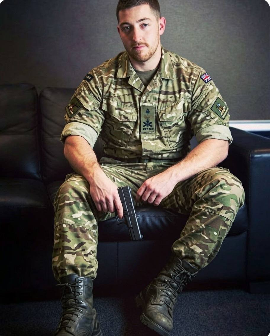 A masculine man in a British military camouflage uniform and combat boots sits on a black leather couch while holding a handgun.