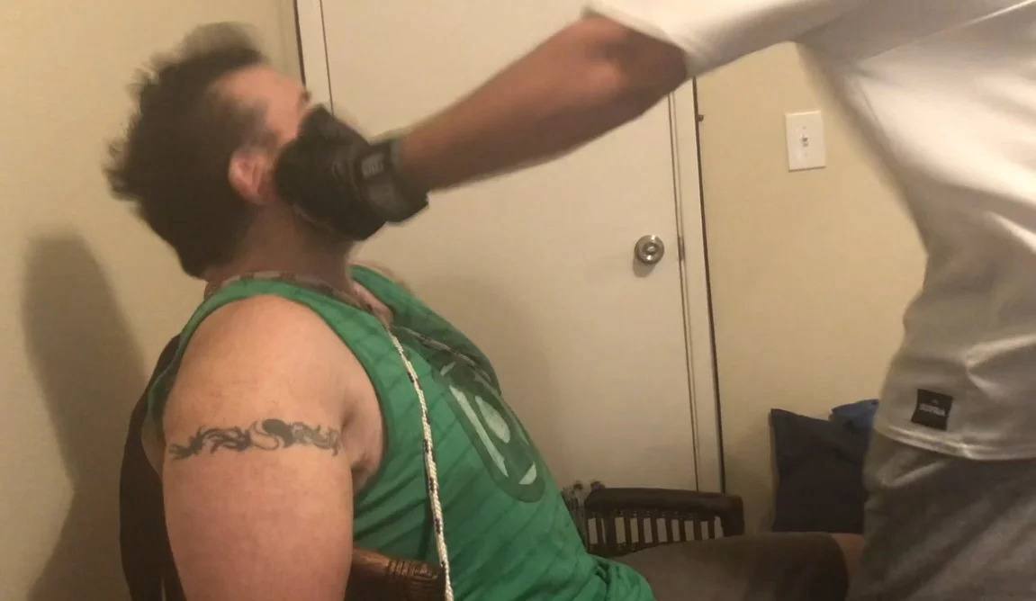A man wearing a white t-shirt strikes a seated man in the face with a gloved hand indoors.