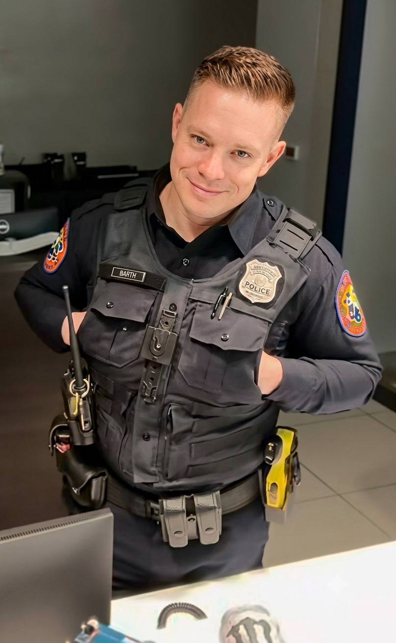 A man wearing a complete police uniform with a tactical vest and utility belt stands in an office setting.
