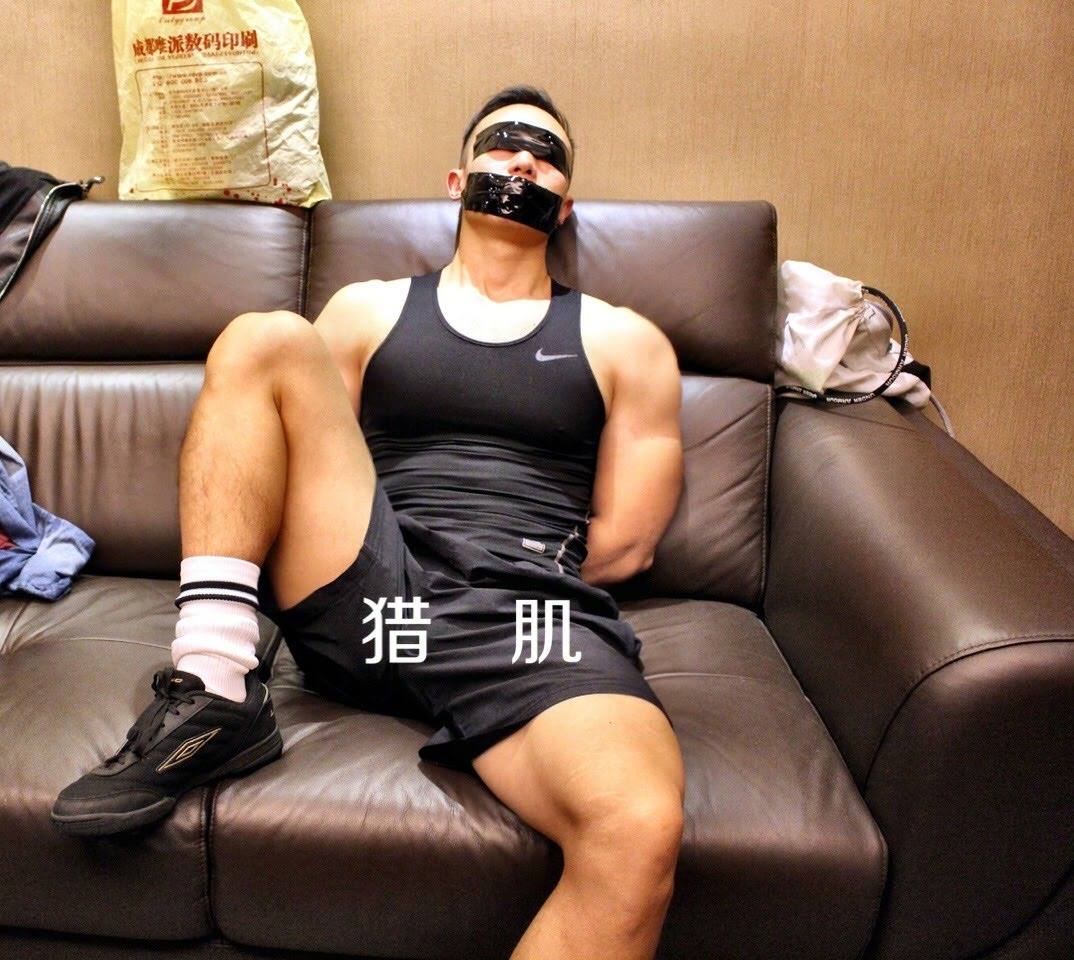 A muscular man wearing sports gear sits restrained on a brown leather couch with black duct tape covering his eyes and mouth.