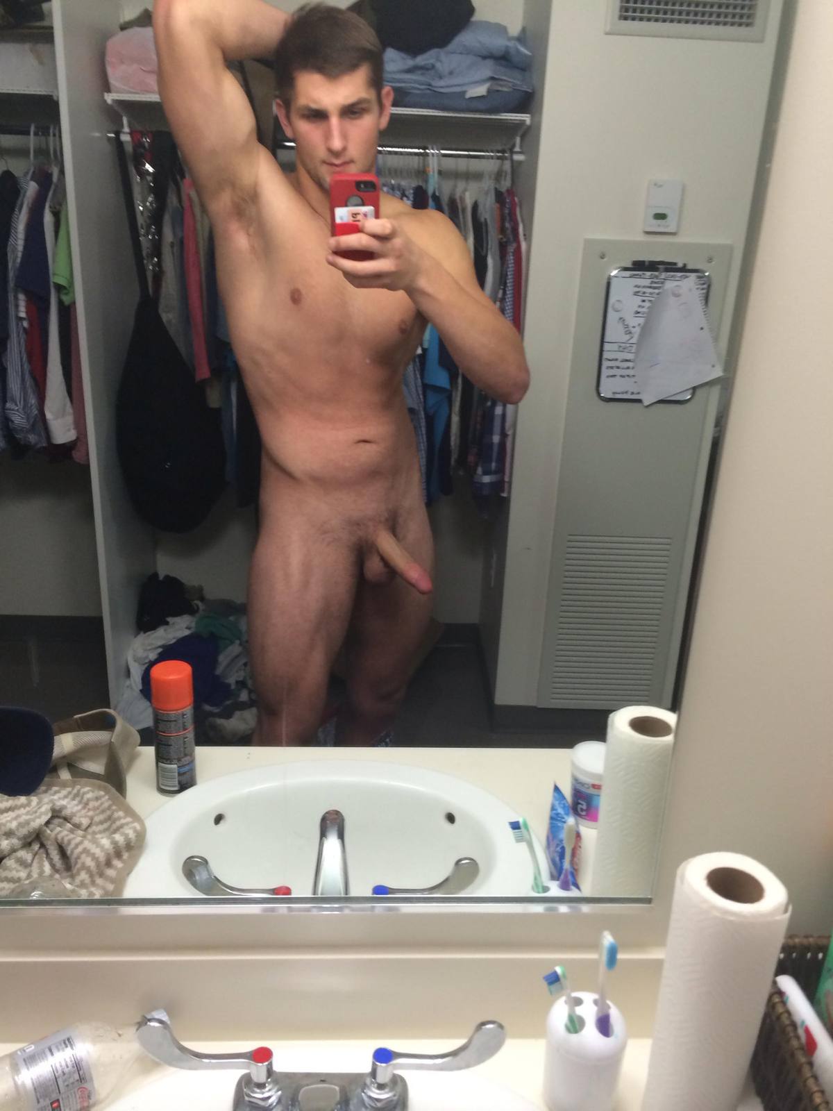A fit young man takes a full-body nude mirror selfie inside a bathroom.
