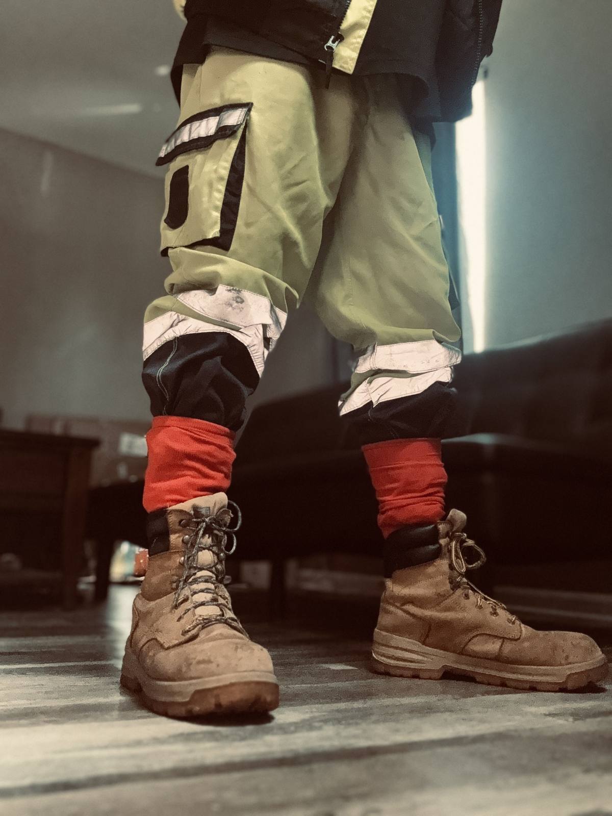 A man wears high-visibility construction pants tucked into red socks and tan work boots in an indoor setting.