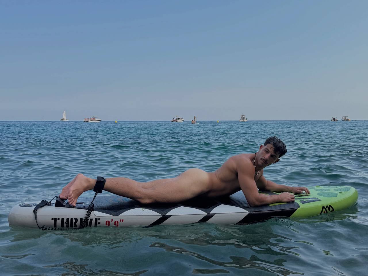 A muscular naked man lies prone on a paddleboard in the open ocean with boats in the background.