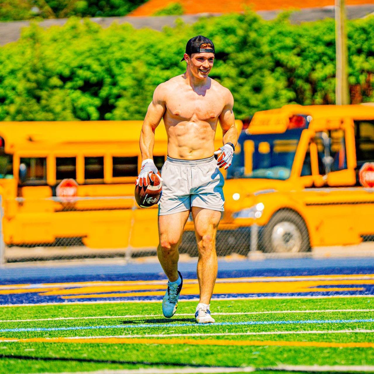 A shirtless, muscular jock in gym shorts and a baseball cap walks across a football field holding a ball.