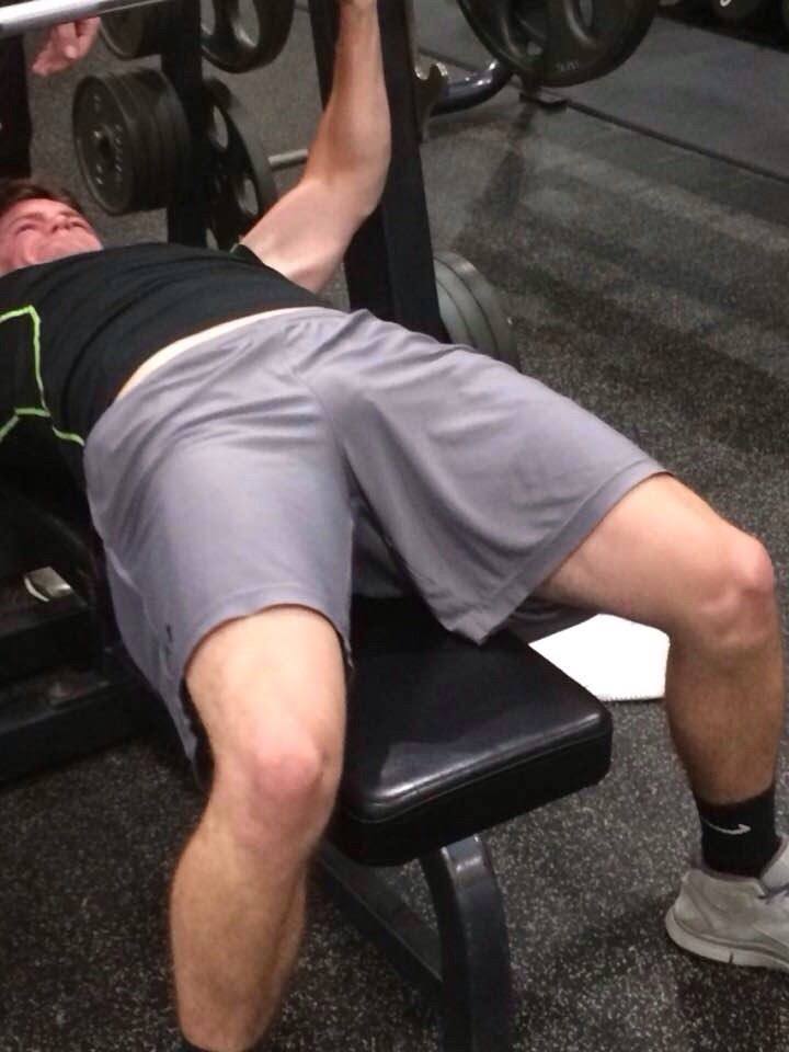 A muscular man in grey gym shorts lies on a weight bench, showing a visible print through his clothing.
