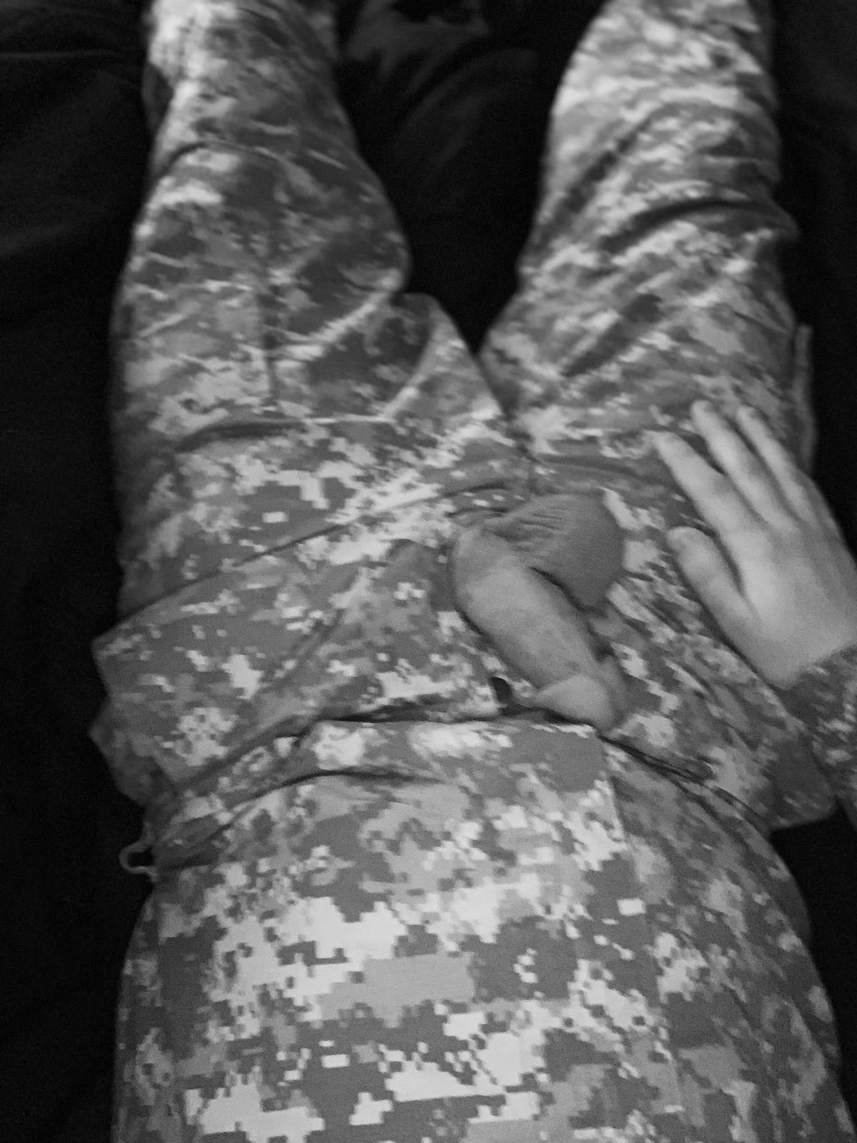 A man in a digital camouflage military uniform lies back with his penis exposed through his trousers.
