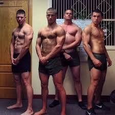 Four shirtless, muscular jocks in gym shorts pose together indoors, showcasing their physiques.