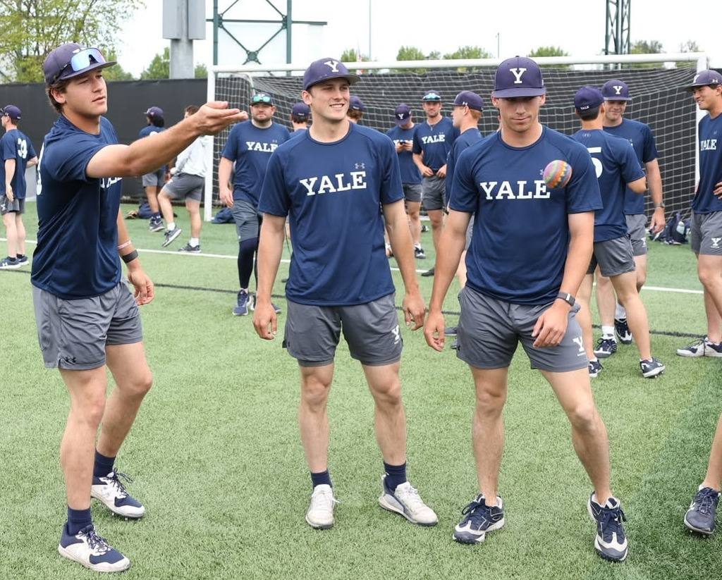 A group of athletic young men in matching Yale university sports gear stand on a turf field during a training session.