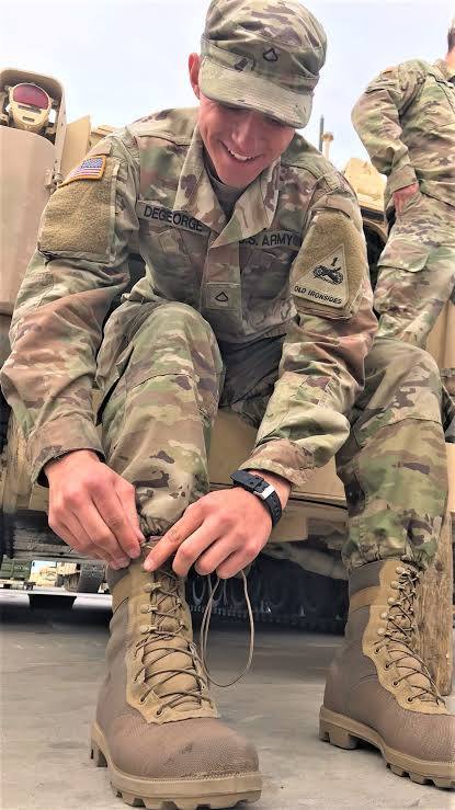 A smiling young man in a full camouflaged US Army uniform sits on a military vehicle while tying the laces of his tan combat boots.