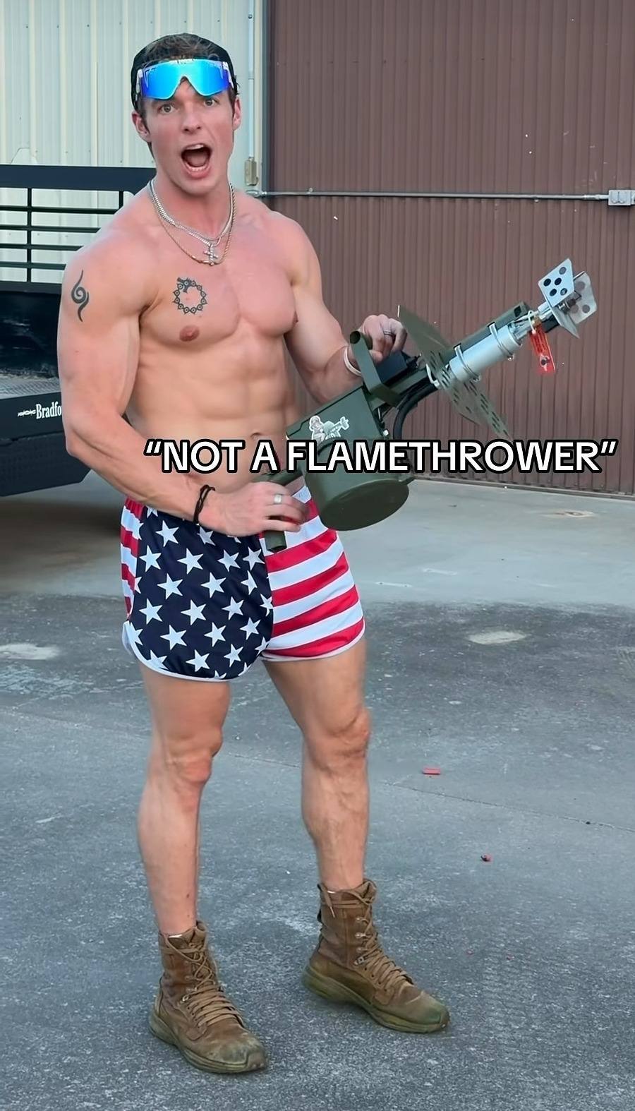 A shirtless muscular man in American flag gym shorts and combat boots poses outdoors holding a flamethrower prop.