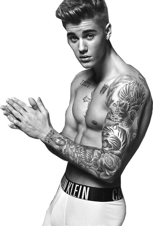 A black and white studio portrait of a shirtless twunk with extensive arm and chest tattoos wearing white branded boxer briefs.