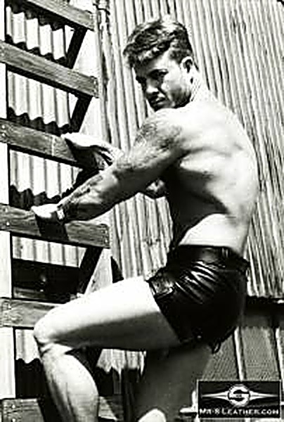 A shirtless muscular man wearing black leather shorts poses on a wooden ladder outdoors against a corrugated metal wall.