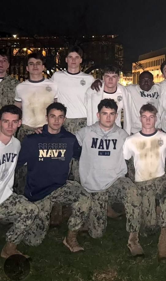 A group of young men in US Navy uniforms, hoodies, and camo pants pose together outdoors at night, with several wearing dirt-stained white t-shirts.