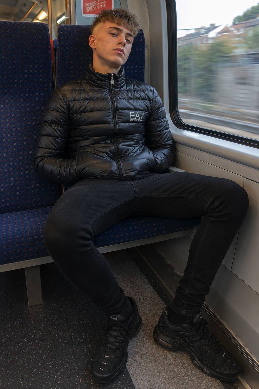 A young man in a black puffer jacket and skinny jeans sits with legs apart on a train seat.