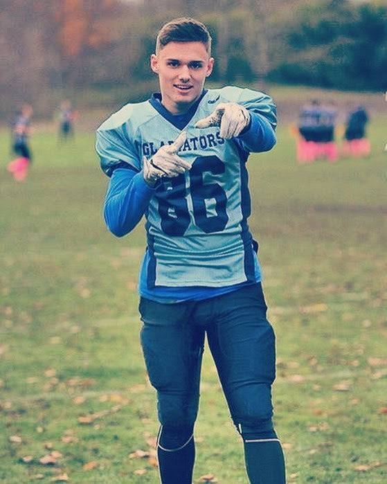 A young athletic man poses outdoors on a grassy field wearing a complete blue football kit with padded pants and gloves.