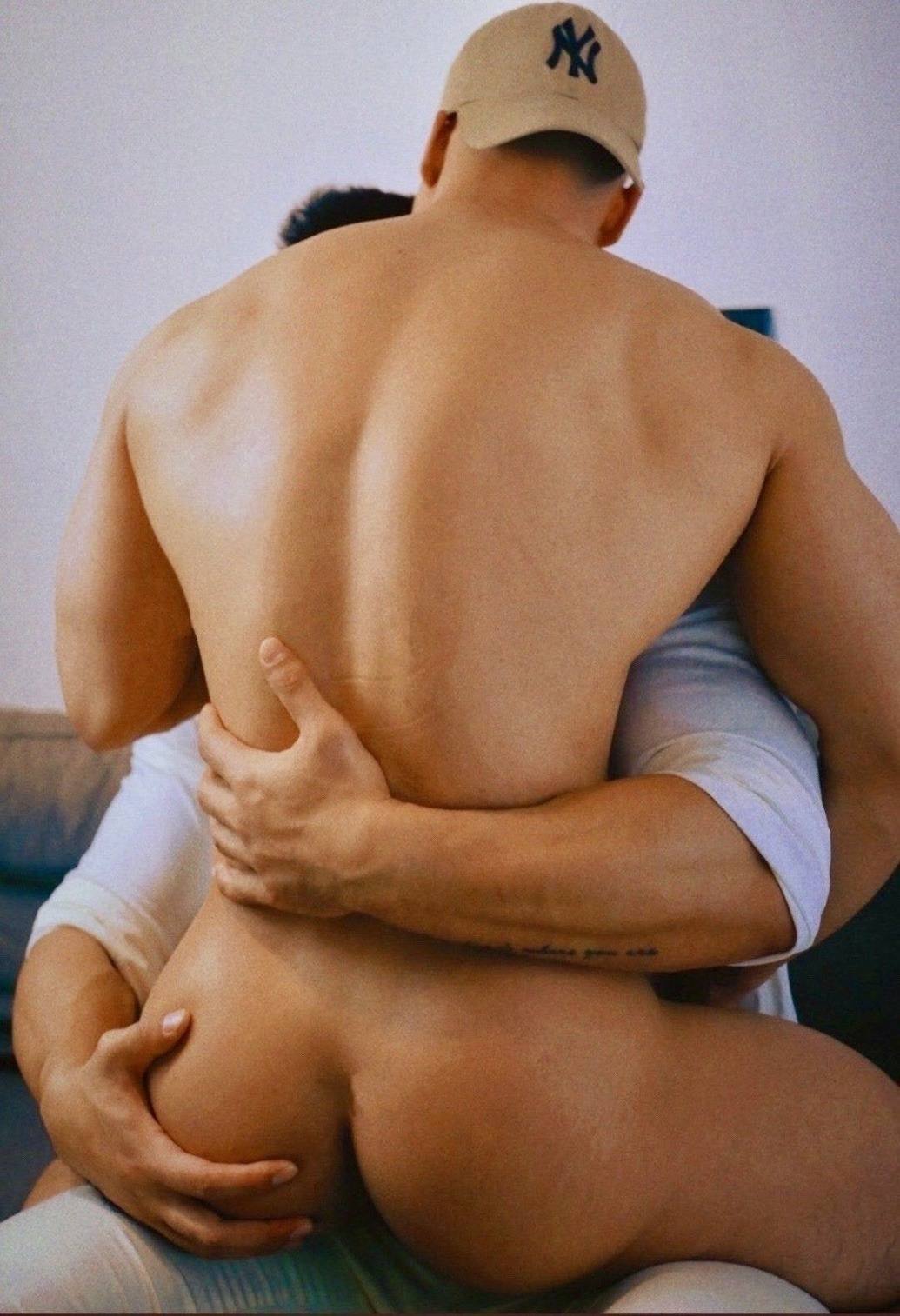 A muscular shirtless man wearing a baseball cap is held from behind by another man, showcasing his bare buttocks and broad back.