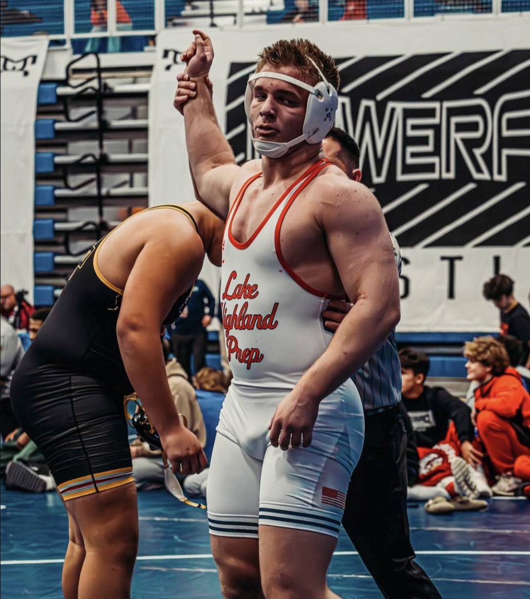 A muscular wrestler in a white and red spandex singlet has his arm raised in victory on the mat.