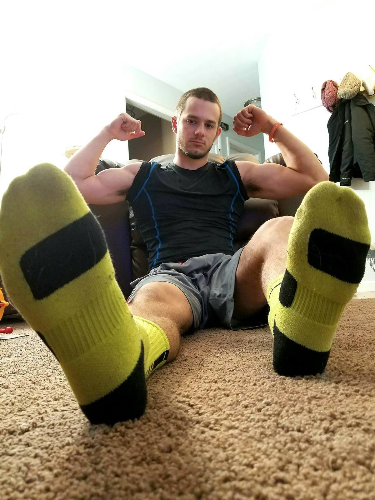 A muscular man in athletic wear sits on the carpet with his socked feet extended toward the camera while flexing his biceps.