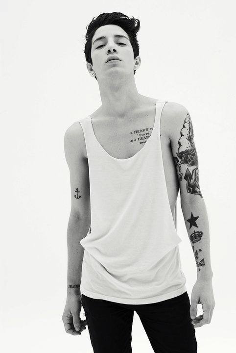 A slim, tattooed twink poses in a deep-cut white tank top and black jeans in a black and white studio shot.