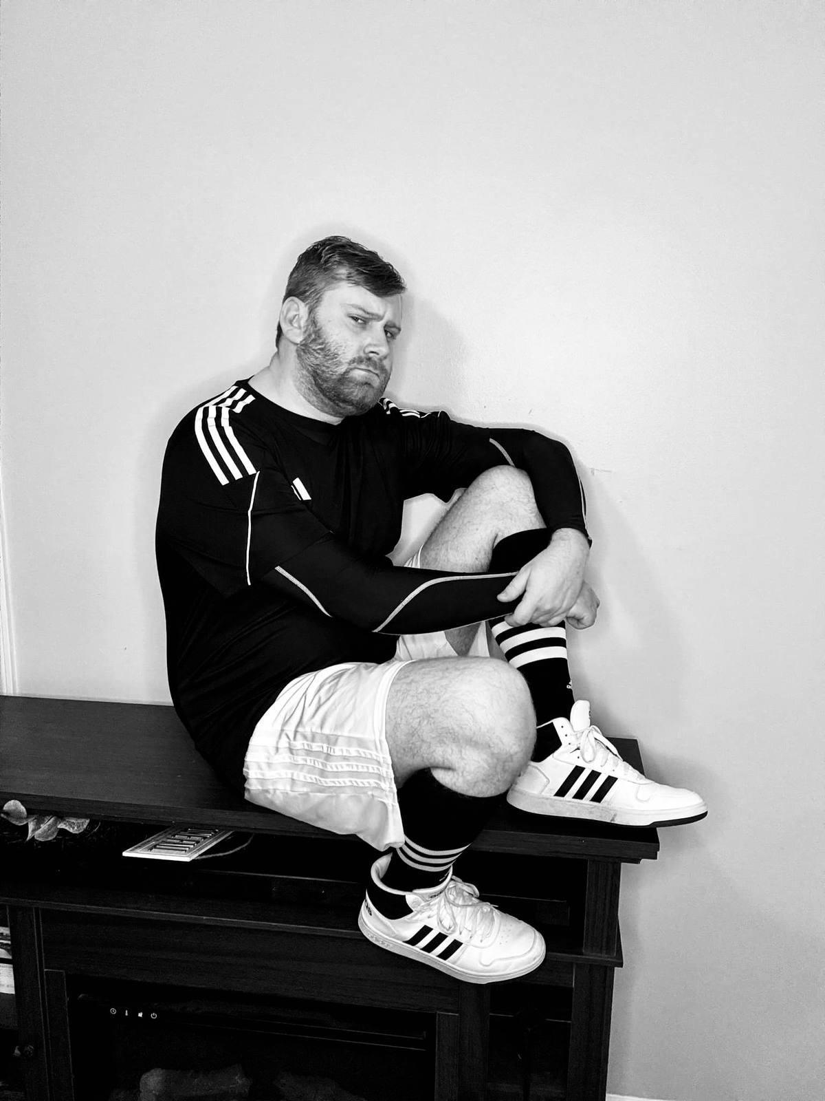 A bearded man in a full athletic kit including a compression shirt, white shorts, and sneakers sits on a wooden ledge.