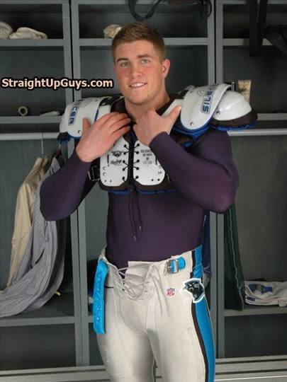 A young athletic man poses in football shoulder pads, a long-sleeve compression shirt, and football pants inside a locker room.