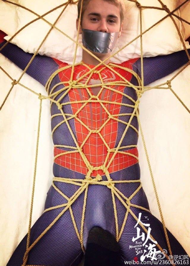 A young man in a Spiderman spandex suit is gagged with duct tape and intricately bound with ropes on a bed.