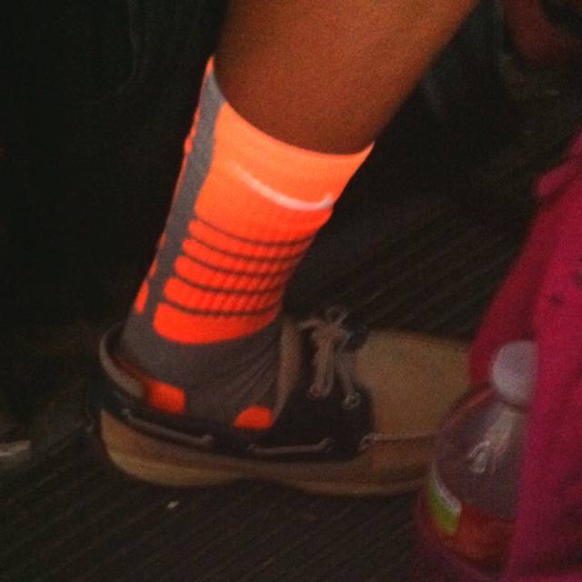 A close-up view of a person's foot wearing a neon orange and grey athletic sock paired with boat shoes in a public setting.