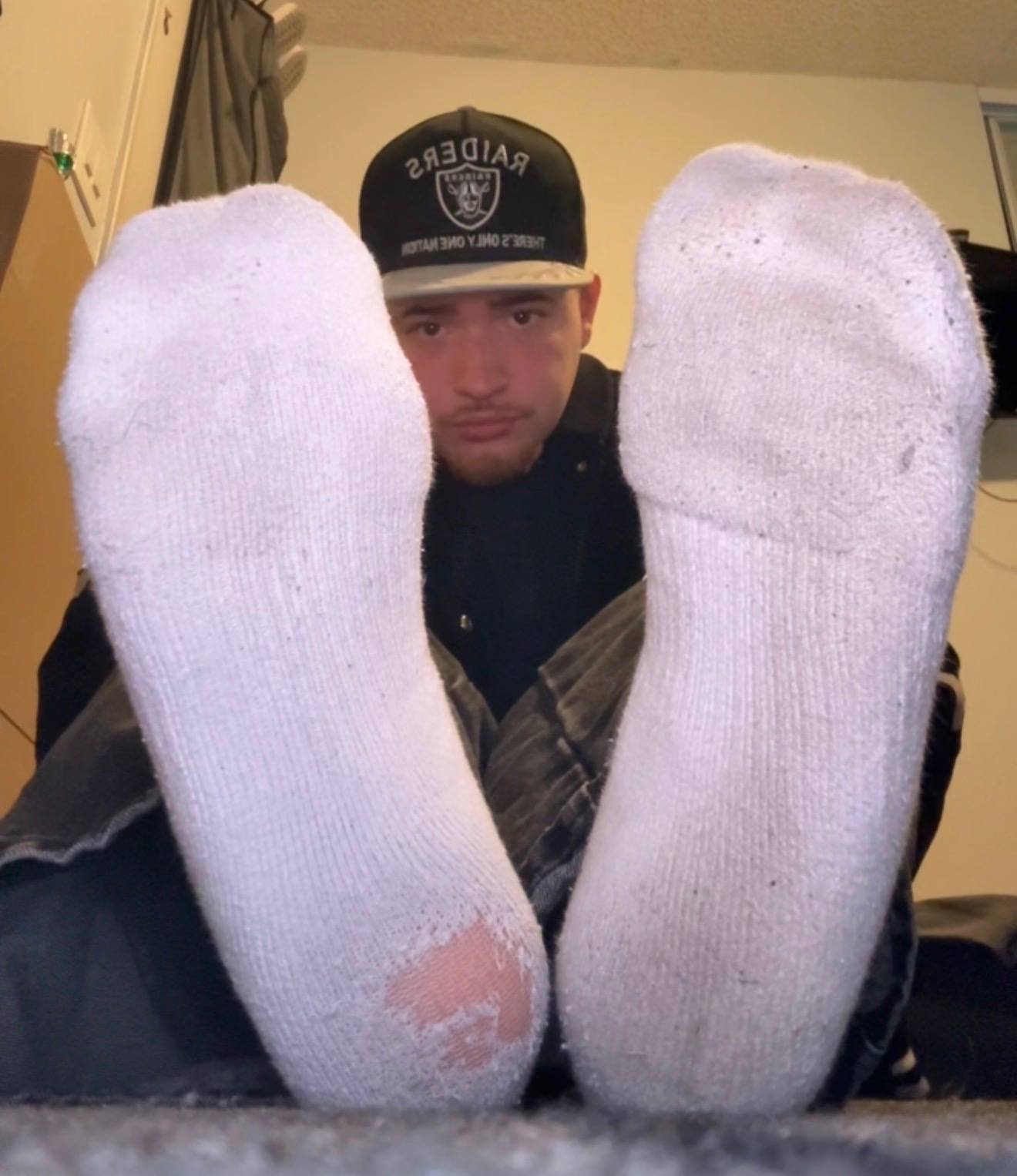 A young man sitting on the floor presents the soles of his worn white socks directly to the camera.