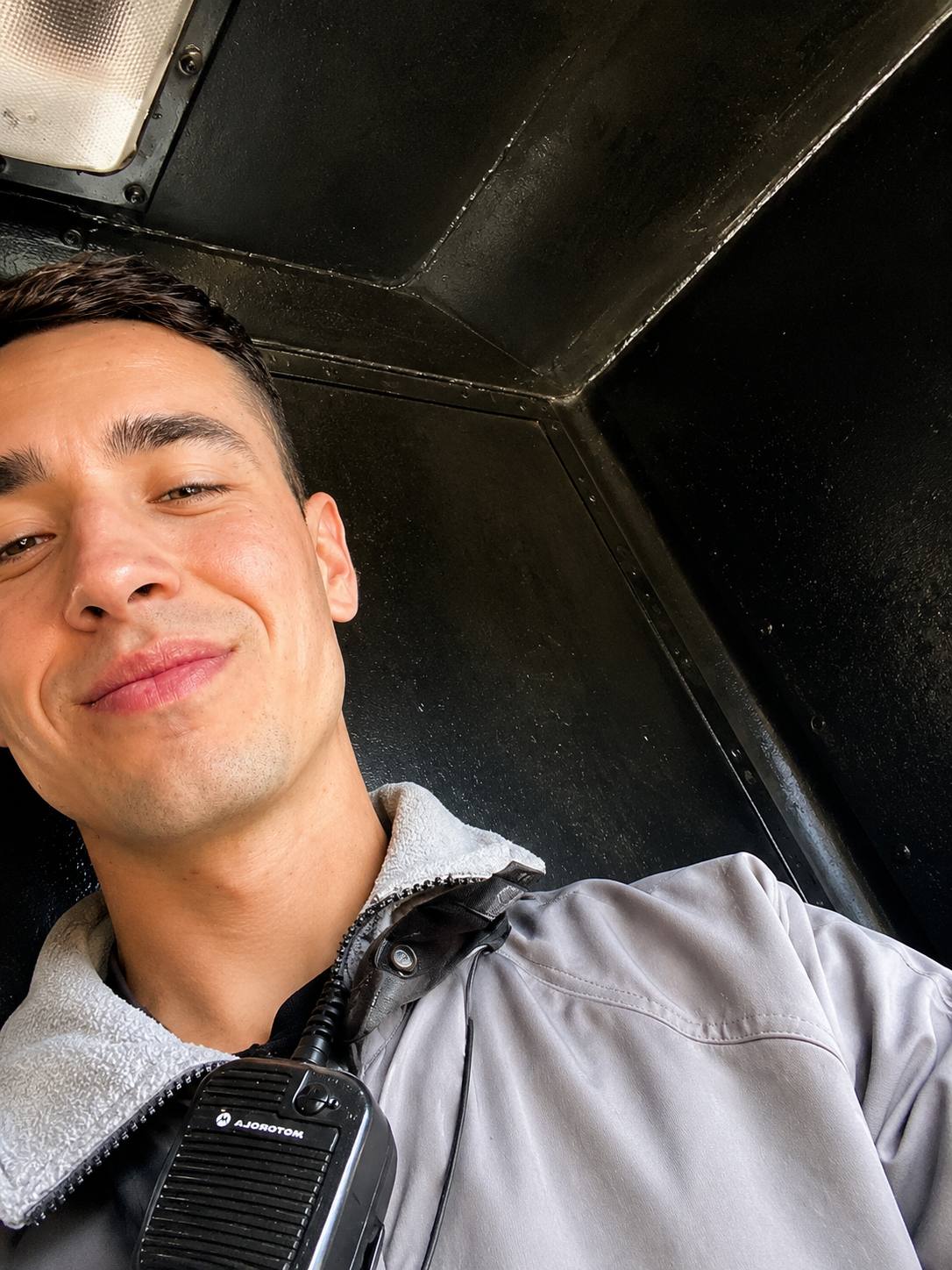 A smiling young man in a grey uniform jacket with a walkie-talkie clipped to his collar poses for a selfie in a dark, enclosed space.