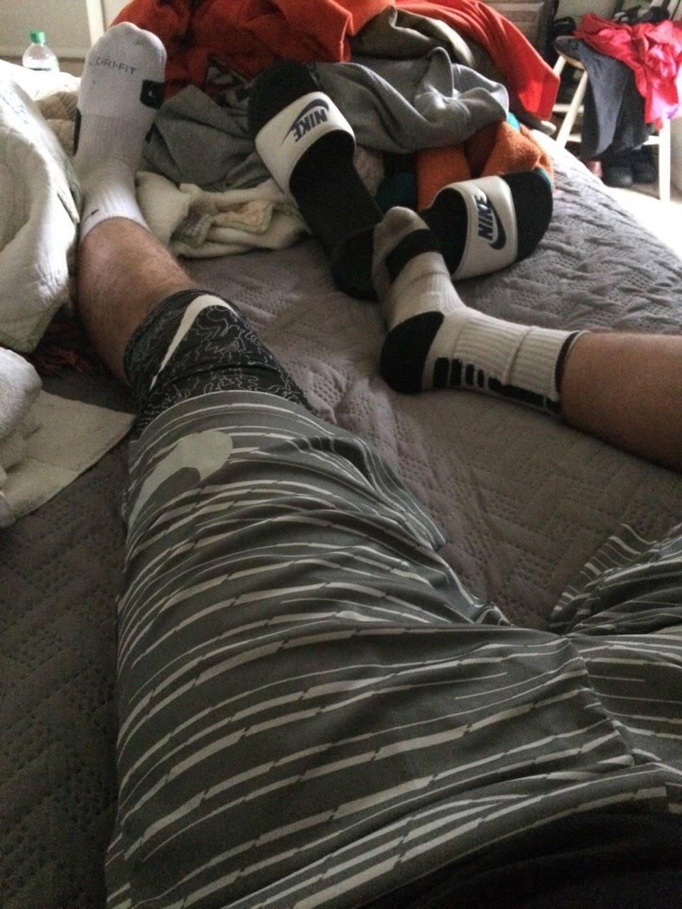 A man with hairy legs relaxes on a bed wearing athletic shorts, crew socks, and Nike slides.