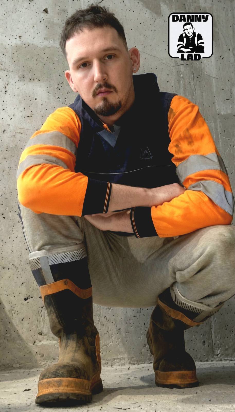 A bearded man in a high-visibility shirt and grey sweatpants squats in front of a concrete wall while wearing muddy work boots.
