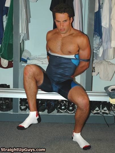 A muscular jock sits on a locker room bench wearing a wrestling singlet pulled down to his waist with his arms behind his back.