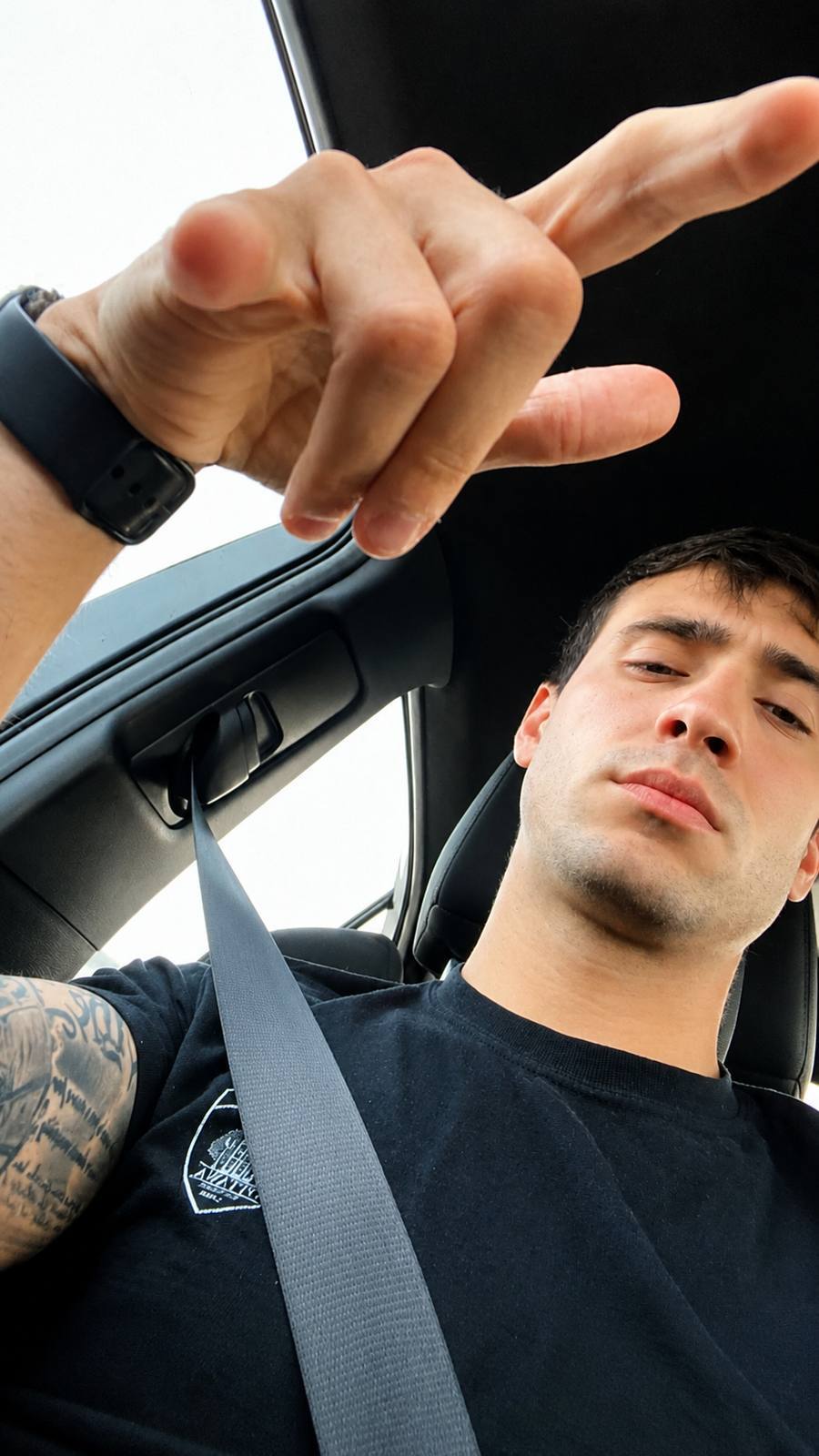 A tattooed young man with short hair and stubble looks down into the lens while sitting in a car.