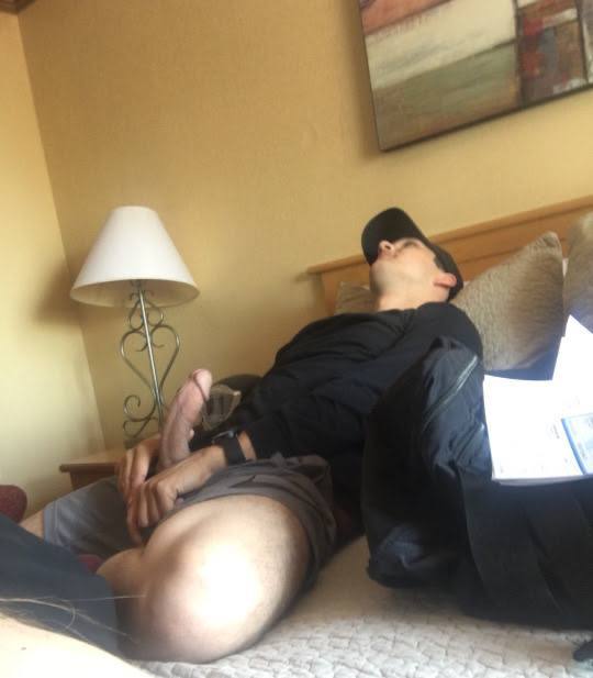 An athletic man wearing a black shirt and baseball cap reclines on a hotel bed exposing his erect penis.