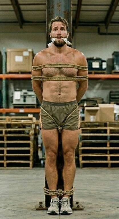 A muscular bearded man is tied to a warehouse support beam with rope around his chest, waist, and ankles while wearing a cloth gag.