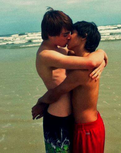 Two young shirtless men in swim trunks embrace and kiss on the beach with ocean waves in the background.