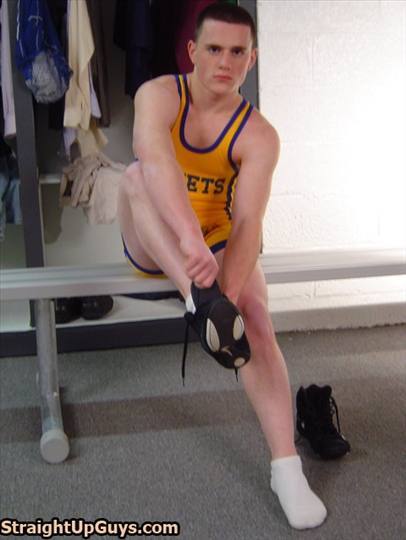 A young man with a buzzcut sits on a locker room bench wearing a yellow wrestling singlet and white socks while putting on his wrestling shoes.
