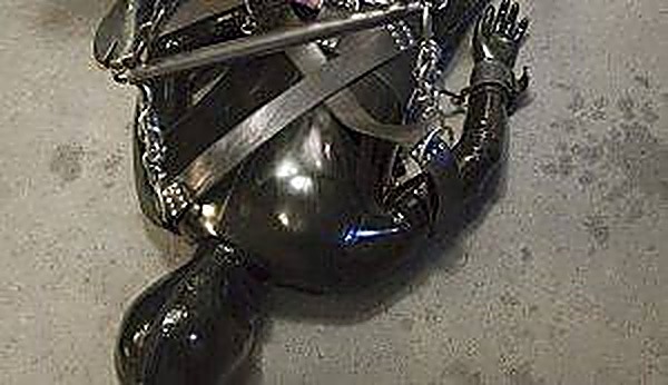 A person fully encased in a shiny black latex suit lies bound on a concrete floor with heavy metal chains and bondage straps.
