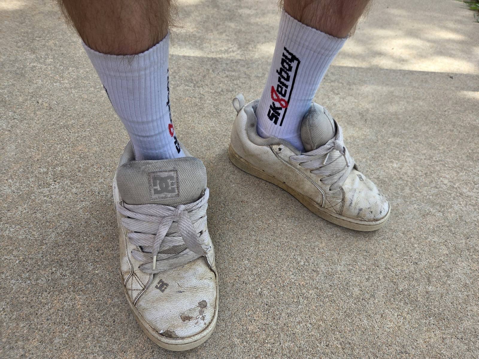 A person stands outdoors wearing white "SK8erboy" socks and dirty white sneakers.