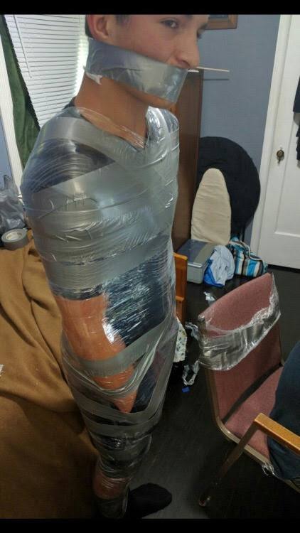 A man stands bound in a domestic setting, his entire body and mouth wrapped in layers of clear plastic film and silver duct tape.