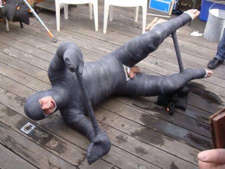 A man lies on a wooden deck, completely immobilized in a grey full-body cast with a cutout exposing his genitals.