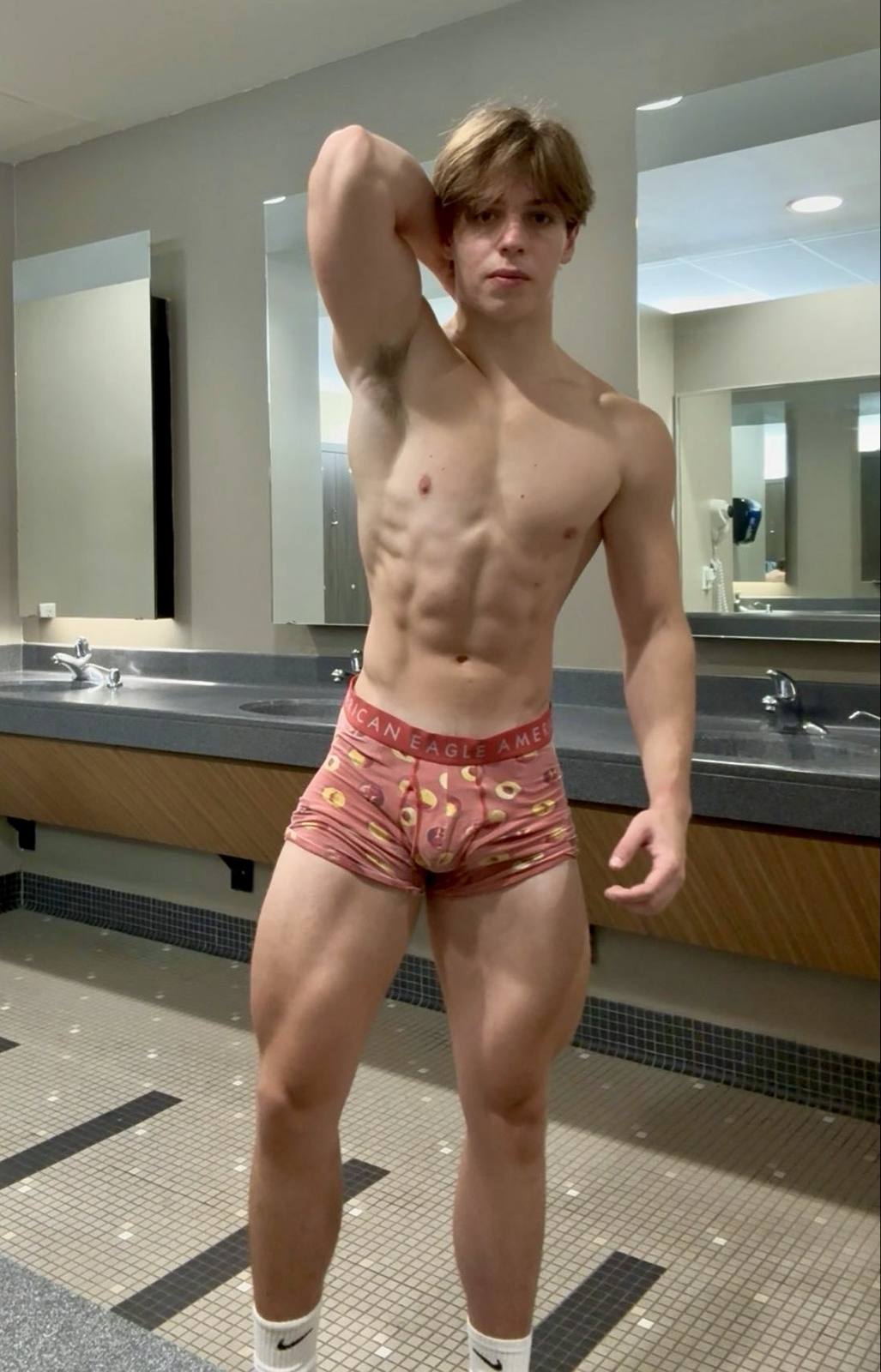 A young muscular man poses in pink patterned boxer briefs and white socks in a public locker room.