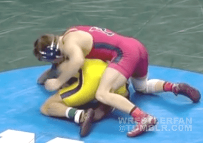 Two male wrestlers in singlets and headgear grapple on a wrestling mat during a match.