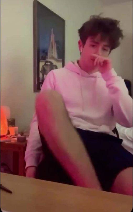 A young man in a pink hoodie and dark shorts sits casually in a room.