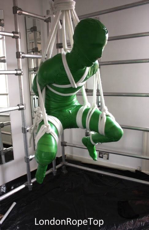 A person wearing a full-body green spandex suit is suspended in mid-air by white rope bondage within a metal dungeon frame.