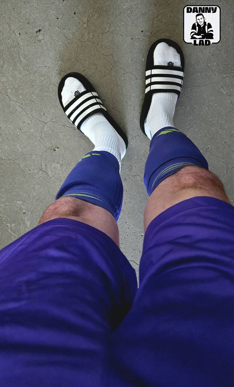 A point-of-view shot of a man with hairy legs wearing purple gym shorts, blue compression sleeves, white tube socks, and striped slides on a concrete floor.