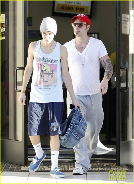 Two men, one young and slim in a graphic tank top and gym shorts, walk out of a venue alongside an older man in sweatpants and a red cap.