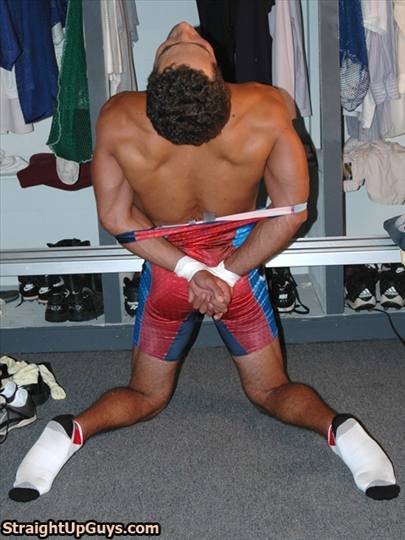 A muscular man in a red and blue wrestling singlet is kneeling in a locker room with his hands bound behind his back.