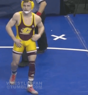 A muscular young wrestler in a yellow and maroon singlet adjusts his gear while standing on a blue wrestling mat.